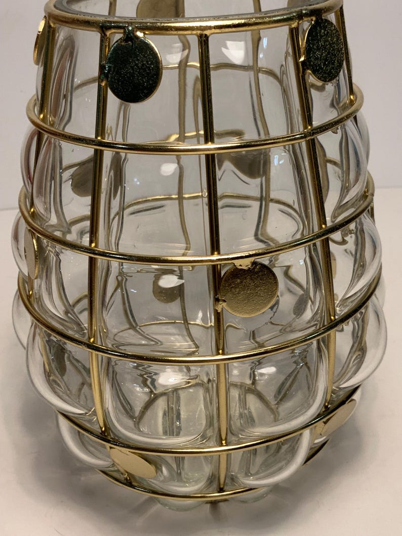 Op de afbeelding: Een heldere glazen vaas met een bubbelachtige textuur, omhuld door een gouden metalen kooi. De kooi heeft cirkelvormige gouden accenten. De vaas heeft een uniek, decoratief ontwerp, geschikt voor het tentoonstellen van bloemen of als een op zichzelf staand kunstwerk.