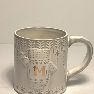 May include: A white ceramic mug with a gray mitten design and the gold letter "M". The mug has a gray handle and is decorated with snowflakes and leaf patterns. A cozy winter-themed mug.
