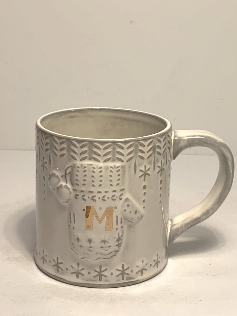 May include: A white ceramic mug with a gray mitten design and the gold letter "M". The mug has a gray handle and is decorated with snowflakes and leaf patterns. A cozy winter-themed mug.