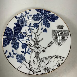 May include: A white porcelain plate with a blue and black design. The plate features a deer with a crown and a coat of arms. The design is reminiscent of a vintage pattern.
