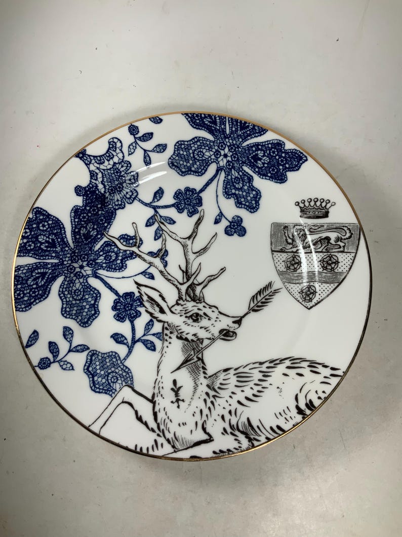 May include: A white porcelain plate with a blue and black design. The plate features a deer with a crown and a coat of arms. The design is reminiscent of a vintage pattern.