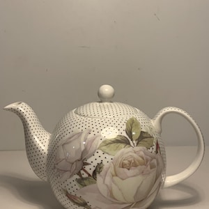 Può includere: Teiera in ceramica bianca con motivo a pois neri e design floreale di rose. La teiera ha un beccuccio curvo, un manico arrotondato e un piccolo pomello sul coperchio. Le rose sono nelle tonalità del bianco e del crema con foglie verdi.