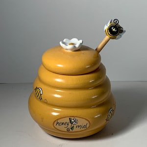 Puede incluir: Un tarro de miel de cerámica amarilla con forma de colmena, con una flor blanca en la tapa. El tarro está decorado con abejas de dibujos animados y tiene una etiqueta que dice "honey miel". Incluye un cucharón de miel de madera con una abeja encima.