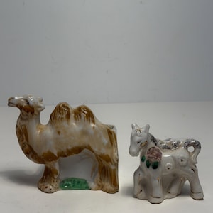Puede incluir: Dos figuras de cerámica: un camello y un caballo. El camello es marrón y blanco con detalles marrones y base verde. El caballo es blanco con detalles grises y dorados, y un diseño floral rosa y verde.