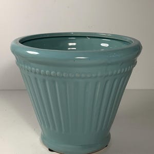 May include: A teal ceramic planter with a fluted design and a beaded trim. The planter has a wide opening and a slightly flared base. The color is a soft, muted teal, suitable for indoor or outdoor use.