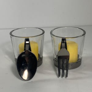 May include: Two clear glass votive candle holders with yellow candles inside. One holder has a spoon-shaped handle, the other a fork-shaped handle. The holders have a silver-colored metal base.