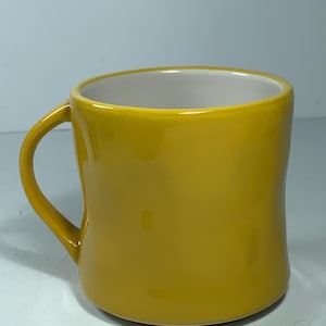 Puede incluir: Una taza de cer&aacute;mica amarilla mostaza con un asa curva. La taza tiene un acabado liso y brillante y un interior blanco. La taza es de color liso y parece estar vac&iacute;a.
