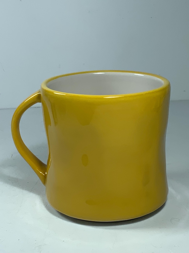 Puede incluir: Una taza de cer&aacute;mica amarilla mostaza con un asa curva. La taza tiene un acabado liso y brillante y un interior blanco. La taza es de color liso y parece estar vac&iacute;a.