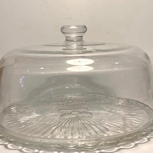 May include: A clear glass cake stand with a domed lid. The base has a scalloped edge and a floral design in the center. The lid has a small knob on top for easy handling. The stand is designed to display and protect baked goods.