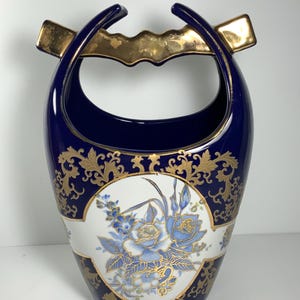 May include: A blue and gold ceramic vase with a floral design. The vase has a unique handle that is shaped like a loop. The vase is decorated with gold accents and a white floral design.