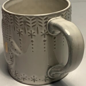 May include: A ceramic mug with a light gray glaze and a decorative pattern of snowflakes and leaves. The handle and rim have a slightly darker gray hue. The word "MOM" is visible in gold lettering on the side of the mug.