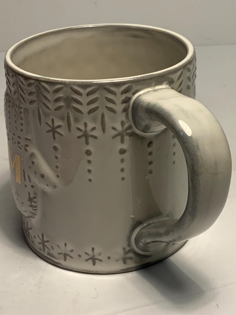 May include: A ceramic mug with a light gray glaze and a decorative pattern of snowflakes and leaves. The handle and rim have a slightly darker gray hue. The word "MOM" is visible in gold lettering on the side of the mug.