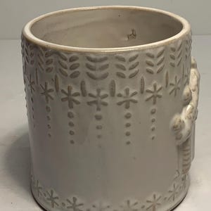 Peut inclure: Pot de fleurs cylindrique en c&eacute;ramique blanc cass&eacute; avec des motifs floraux et &eacute;toil&eacute;s en relief. Le design comprend des rang&eacute;es verticales de feuilles et de points suspendus, avec une bordure d'&eacute;toiles &agrave; la base. Une figure est en relief sur le c&ocirc;t&eacute;.