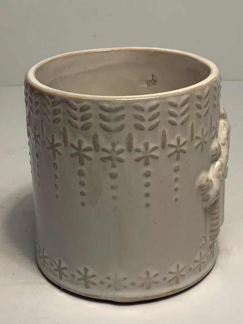 Peut inclure: Pot de fleurs cylindrique en c&eacute;ramique blanc cass&eacute; avec des motifs floraux et &eacute;toil&eacute;s en relief. Le design comprend des rang&eacute;es verticales de feuilles et de points suspendus, avec une bordure d'&eacute;toiles &agrave; la base. Une figure est en relief sur le c&ocirc;t&eacute;.