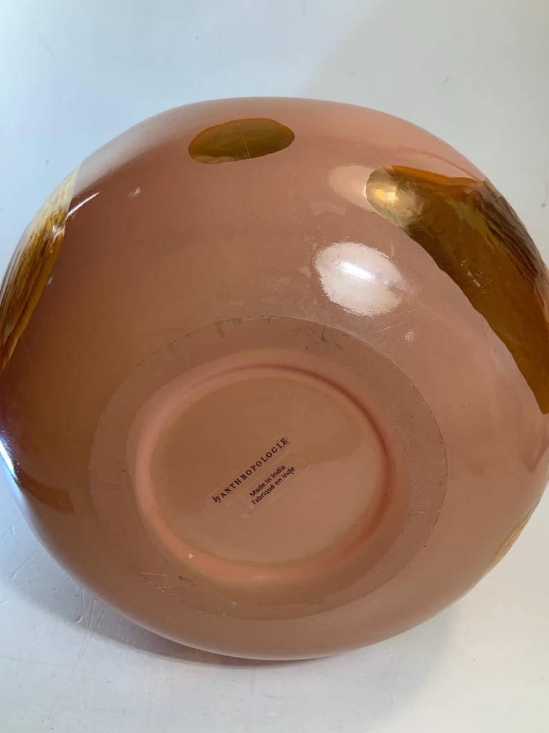 May include: A peach-colored, round decorative bowl with gold accents. The bowl has a wide base and a rounded top. The bottom of the bowl has the text "by ANTHROPOLOGIE Made in India Fabriqu&eacute; en Inde."