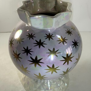 May include: A pearlescent ceramic vase with a spherical body and a star-shaped opening. The vase is decorated with gold star patterns. The base has a geometric design. The vase is approximately 8 inches tall.