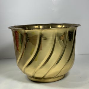 Puede incluir: Un macetero de metal dorado con un diseño de espiral. El macetero es redondo y tiene una abertura ancha.