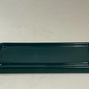 May include: A rectangular, dark teal ceramic tray with a raised rim. The tray has rounded corners and a glossy finish. The interior surface is smooth and reflects light. The tray is designed for holding small items.