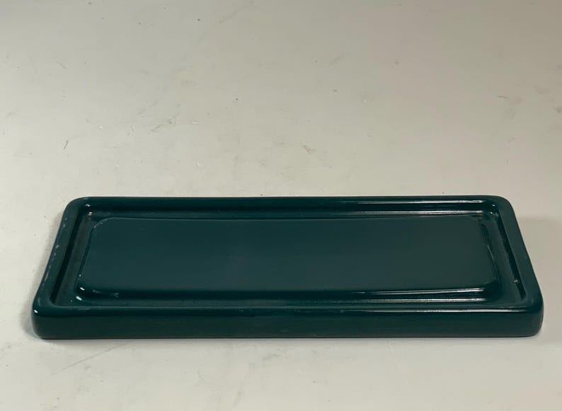 May include: A rectangular, dark teal ceramic tray with a raised rim. The tray has rounded corners and a glossy finish. The interior surface is smooth and reflects light. The tray is designed for holding small items.