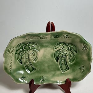 Puede incluir: Una bandeja rectangular de cerámica con borde ondulado e irregular, esmaltada en tonos verdes. La superficie presenta dos diseños de palmeras en relieve y patrones de remolinos decorativos a lo largo del borde, sobre un soporte de madera rojo oscuro.