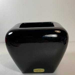 Puede incluir: Un jarrón negro brillante con una abertura cuadrada y un cuerpo redondeado. El jarrón tiene una etiqueta que dice "Haeger Floral" y "Macomb, IL, USA". El jarrón es un objeto decorativo.