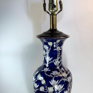 Könnte beinhalten: Blaue Keramik-Tischlampe mit floralem Design. Die Lampe hat einen dunkelblauen Sockel mit weißen Blumen und grünen Blättern. Die Lampe hat einen Messinghals und eine dekorative Spitze. Die Lampe ist etwa 71 cm hoch.