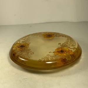 以下が含まれることがあります： 黄色と茶色の花とシダが押し花された、丸い透明なガラス製のコースターです。