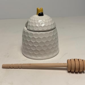 May include: A white ceramic honey pot with a honeycomb pattern and a yellow and black bee-shaped handle. A wooden honey dipper rests beside the pot, ready for use. This kitchenware item is perfect for serving honey.