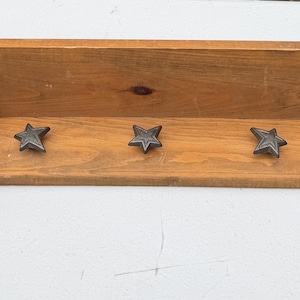 May include: A medium-toned wooden wall shelf featuring a horizontal backboard and a flat base. Three dark, metallic, five-pointed star-shaped hooks are evenly spaced along the front edge of the shelf base, providing a functional design element.