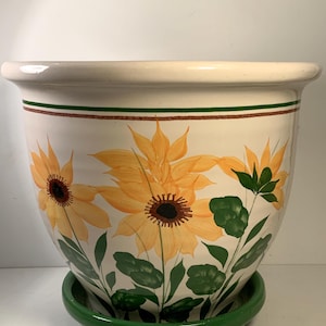 May include: A white ceramic flower pot with a green base, featuring hand-painted yellow sunflowers with brown centers and green leaves. The pot has a brown and green stripe near the rim, perfect for indoor or outdoor plants.