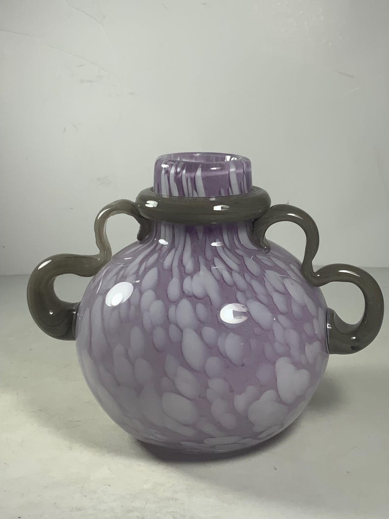 May include: A purple glass vase with white speckles and grey handles. The vase has a round body and a wide opening.
