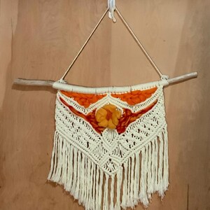 May include: A macrame wall hanging featuring a cream-colored geometric design with orange and yellow accents. The piece is suspended from a natural wooden dowel and includes a fringe at the bottom. A yellow flower is in the center.