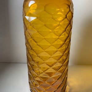 May include: A tall, amber glass bottle with a diamond-shaped pattern. The bottle is empty and has a rounded bottom.
