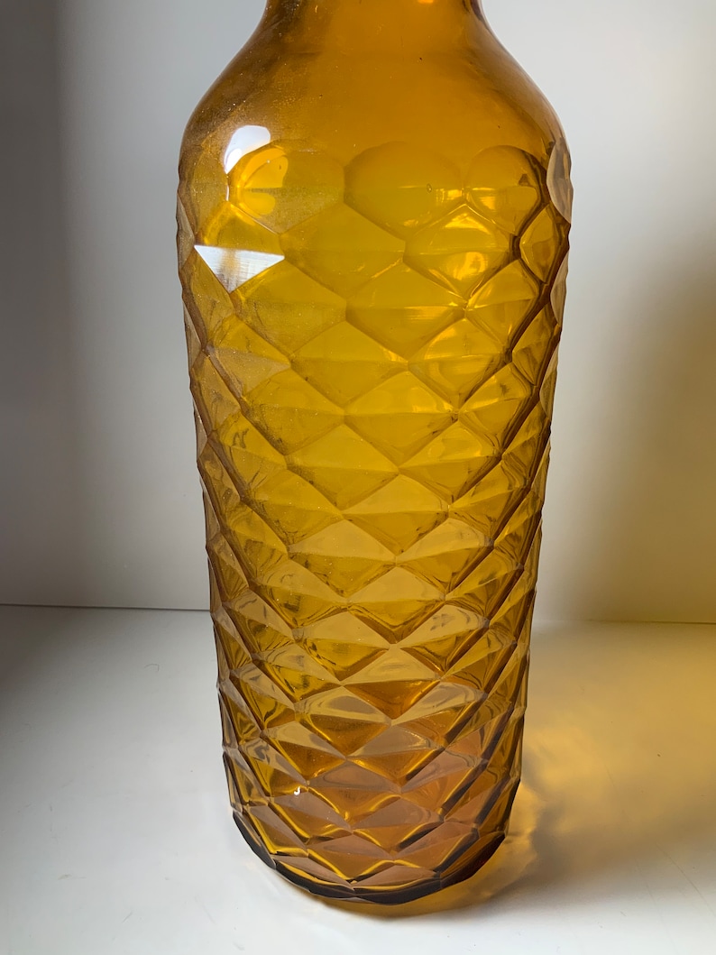 May include: A tall, amber glass bottle with a diamond-shaped pattern. The bottle is empty and has a rounded bottom.