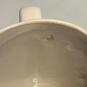 Peut inclure: Gros plan sur une tasse en c&eacute;ramique blanche avec une anse. L'int&eacute;rieur de la tasse est lisse et brillant. Le bord pr&eacute;sente un aspect l&eacute;g&egrave;rement plus fonc&eacute; et vieilli. Un petit &eacute;clat est visible &agrave; l'int&eacute;rieur.