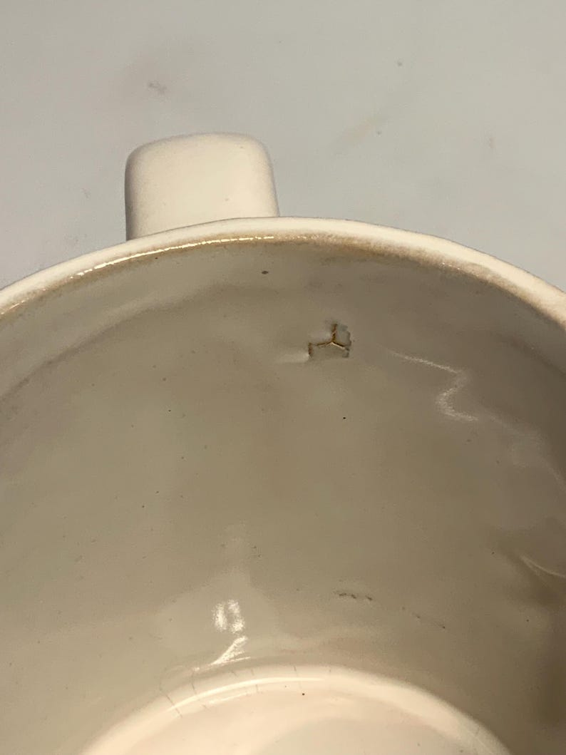 Peut inclure: Gros plan sur une tasse en c&eacute;ramique blanche avec une anse. L'int&eacute;rieur de la tasse est lisse et brillant. Le bord pr&eacute;sente un aspect l&eacute;g&egrave;rement plus fonc&eacute; et vieilli. Un petit &eacute;clat est visible &agrave; l'int&eacute;rieur.