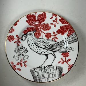 May include: A white ceramic plate with a gold rim features a black and white illustration of a bird perched on a stone structure. The bird is holding a key in its beak. The background is a red floral pattern.