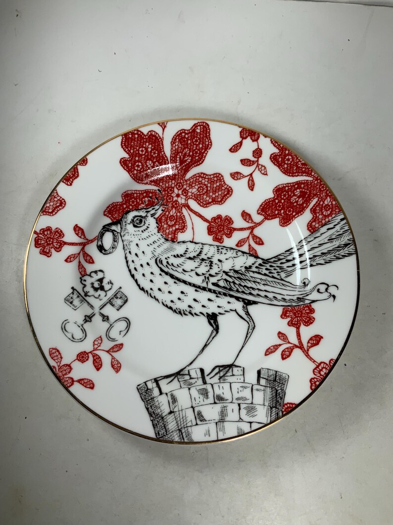 May include: A white ceramic plate with a gold rim features a black and white illustration of a bird perched on a stone structure. The bird is holding a key in its beak. The background is a red floral pattern.