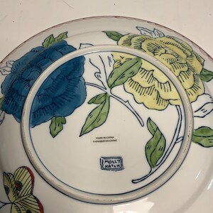 May include: White ceramic plate with a floral design featuring blue and yellow flowers, green leaves, and a red rim. The plate has the text "MADE IN CHINA FABRIQUE EN CHINE" and the "MOLLY HATCH" logo.