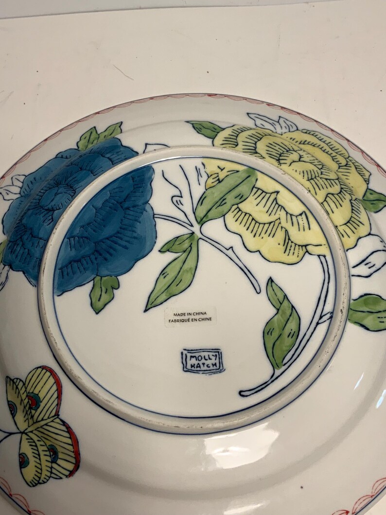 May include: White ceramic plate with a floral design featuring blue and yellow flowers, green leaves, and a red rim. The plate has the text "MADE IN CHINA FABRIQUE EN CHINE" and the "MOLLY HATCH" logo.