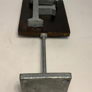 May include: A metal letter "F" sculpture. The letter is mounted on a dark brown rectangular wooden base, supported by a metal rod and a square metal base. The sculpture has a rustic, industrial aesthetic.