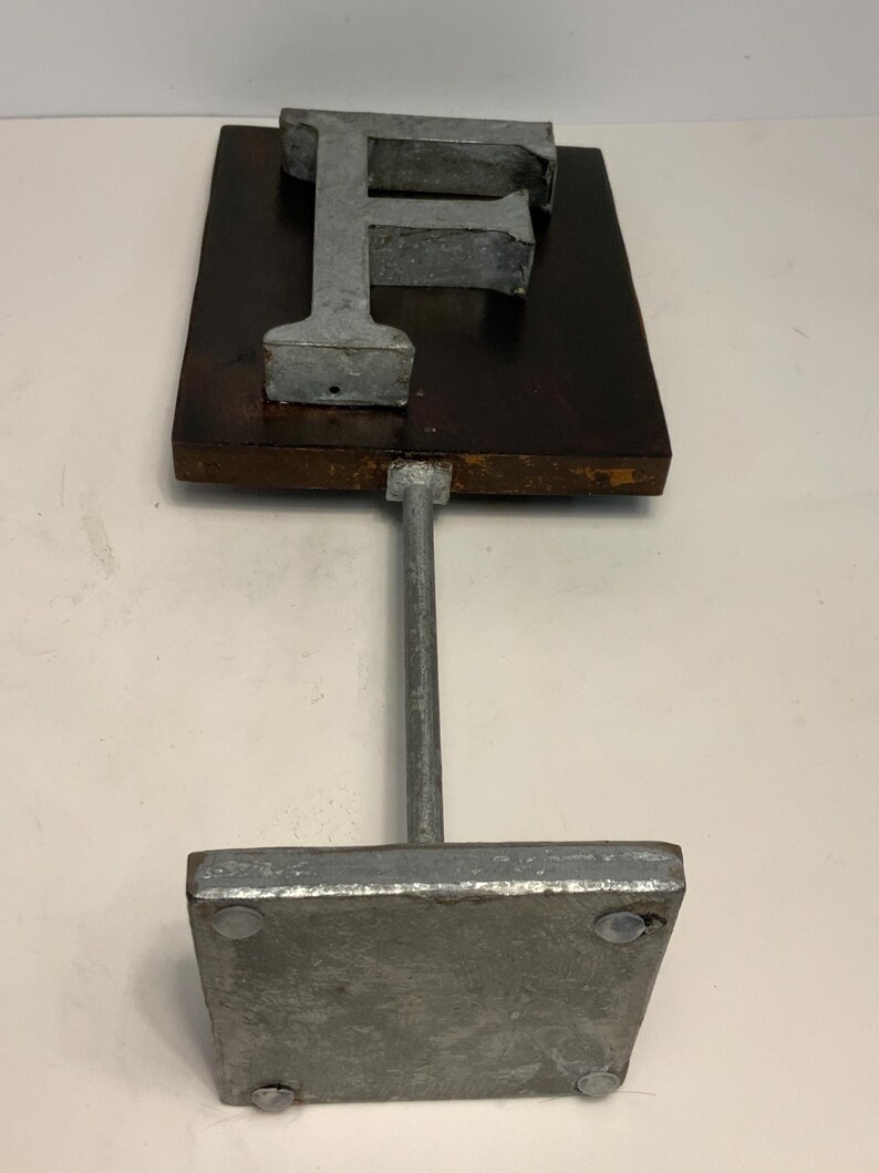 May include: A metal letter "F" sculpture. The letter is mounted on a dark brown rectangular wooden base, supported by a metal rod and a square metal base. The sculpture has a rustic, industrial aesthetic.