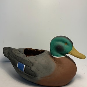 May include: A ceramic duck planter with a green head, brown body, and gray wings. The duck has a yellow beak and a blue accent on its wing. The planter has a hollowed-out area on its back for plants or other decorative items.