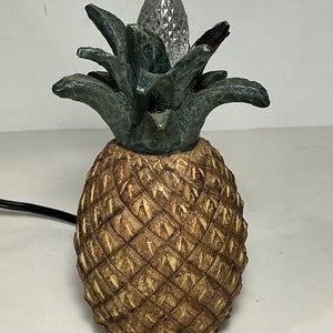 May include: A pineapple-shaped table lamp featuring a textured, golden-brown body with a cross-hatched pattern and a dark green, leafy top. A clear, faceted, flame-shaped light bulb sits at the centre of the green leaves, connected by a black cord.