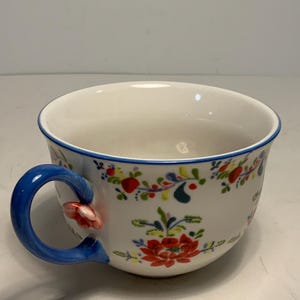 May include: White ceramic teacup with a blue handle and rim, adorned with a floral pattern in red, green, and yellow. The handle features a small red flower accent.