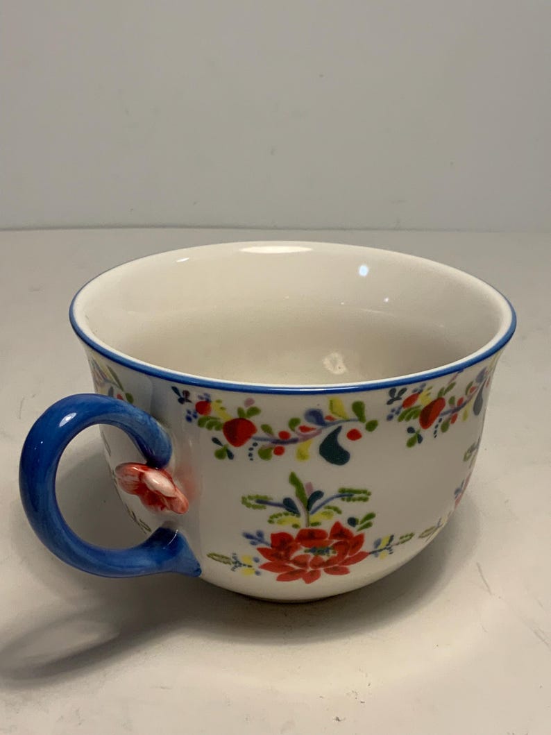 May include: White ceramic teacup with a blue handle and rim, adorned with a floral pattern in red, green, and yellow. The handle features a small red flower accent.