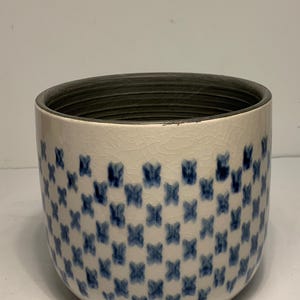 Può includere: Un vaso di ceramica con base bianca e un motivo di disegni blu a forma di croce. Il bordo è grigio scuro e anche l'interno è grigio scuro. Il vaso ha una superficie screpolata.