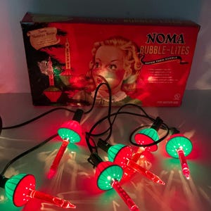 Puede incluir: Un juego de luces de cadena Noma Bubble-Lites con bases de plástico rojo y verde y tubos de vidrio transparente. La caja de cartón roja original muestra el texto Noma Bubble-Lites Watch Them Bubble y una ilustración de una persona.