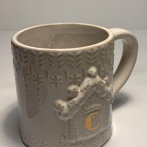 Peut inclure: Une tasse en c&eacute;ramique cr&egrave;me avec une anse. La tasse pr&eacute;sente un motif en relief d'une maison en pain d'&eacute;pices avec une lettre "C" dor&eacute;e sur le c&ocirc;t&eacute;. La tasse a une capacit&eacute; d'environ 350 ml.