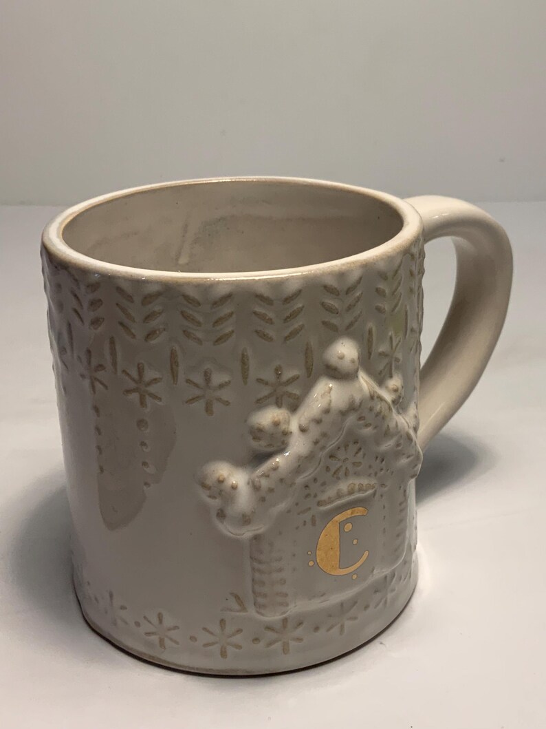 Peut inclure: Une tasse en c&eacute;ramique cr&egrave;me avec une anse. La tasse pr&eacute;sente un motif en relief d'une maison en pain d'&eacute;pices avec une lettre "C" dor&eacute;e sur le c&ocirc;t&eacute;. La tasse a une capacit&eacute; d'environ 350 ml.